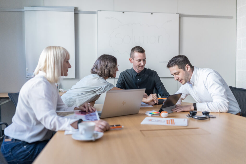 4 Personen sitzen gemeinsam an einem Bürotisch und unterhalten sich über ein Thema. Eine Person zeigt den anderen etwas auf dem Ipad. Es liegen Blöcke, Stifte etc. auf dem Tisch.