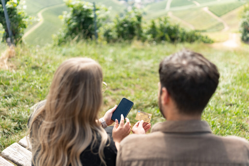 Zwei Personen sitzen gemeinsam in der Natur und geben in ein Smartphone die Bezahldaten einer Kreditkarte ein.